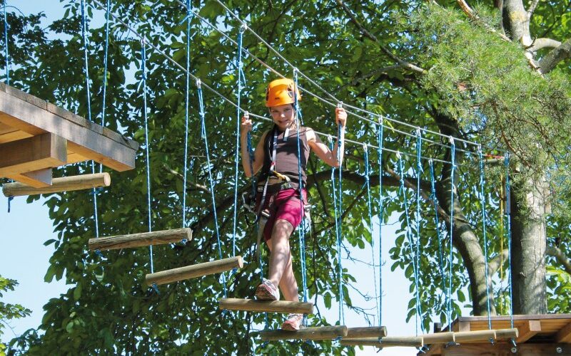 Eine Person, die einen Helm und ein Gurtzeug trägt, überquert eine Seilbrücke in einem von grünen Bäumen umgebenen Outdoor-Abenteuerpark.