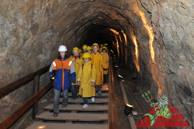 Eine Gruppe Kinder in gelben Regenmänteln und Helmen geht in einem felsigen unterirdischen Tunnel Holzstufen hinunter, angeführt von einem Erwachsenen in einer blau-orangen Jacke.