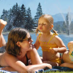 Eine vierköpfige Familie entspannt sich an einem sonnigen Tag auf einer Decke an einem See, im Hintergrund sind Bäume, Berge und rote Sonnenschirme zu sehen.