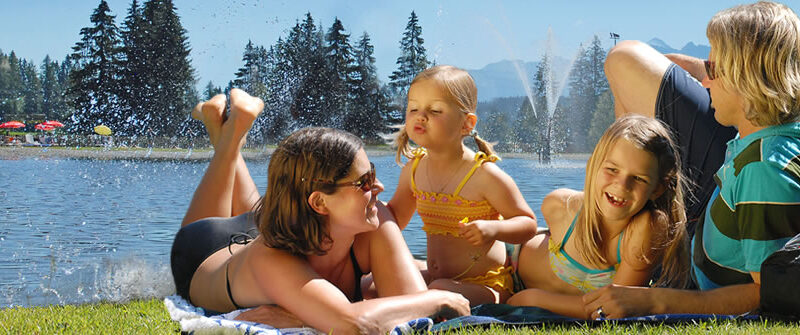 Eine vierköpfige Familie entspannt sich an einem sonnigen Tag auf einer Decke an einem See, im Hintergrund sind Bäume, Berge und rote Sonnenschirme zu sehen.