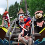Eine Gruppe von als Piraten verkleideten Personen, darunter eine Person in einem Bärenkostüm, posiert auf einem hölzernen Spielschiff im Freien, umgeben von Bäumen.