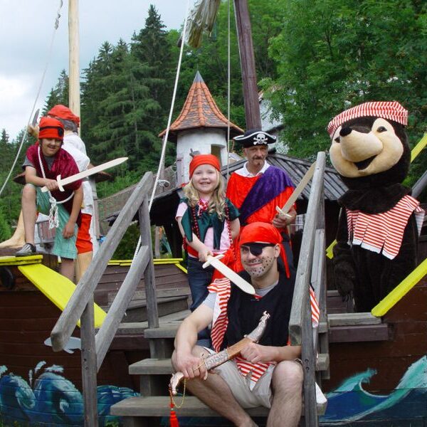 Eine Gruppe von als Piraten verkleideten Personen, darunter eine Person in einem Bärenkostüm, posiert auf einem hölzernen Spielschiff im Freien, umgeben von Bäumen.