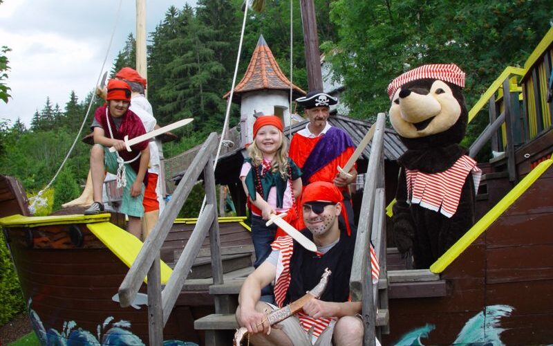 Eine Gruppe von als Piraten verkleideten Personen, darunter ein Pirat im Bärenkostüm, posiert auf einem hölzernen Spielschiff im Freien.