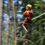 Ein Kind, das einen Helm und ein Gurtzeug trägt, fährt mit einer Seilrutsche durch ein Waldgebiet, wobei die Bäume im Hintergrund verschwimmen.