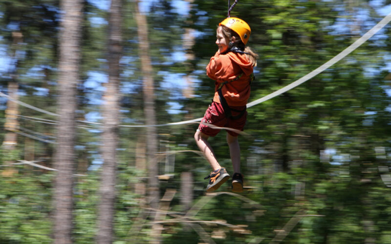 Ein Kind, das einen Helm und ein Gurtzeug trägt, fährt mit einer Seilrutsche durch ein Waldgebiet, wobei die Bäume im Hintergrund verschwimmen.