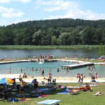 An einem sonnigen Tag entspannen sich die Menschen an einem Strand und Freibad am See, umgeben von Gras, Bäumen und bewaldeten Hügeln im Hintergrund.