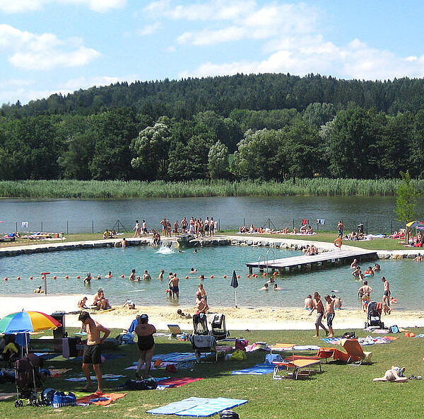 An einem sonnigen Tag entspannen sich die Menschen an einem Strand und Freibad am See, umgeben von Gras, Bäumen und bewaldeten Hügeln im Hintergrund.
