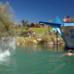 Die Leute schwimmen und sehen zu, wie jemand von einer blauen Holzkonstruktion in Form eines Schiffes in einen See springt und dabei einen großen Sprung macht.