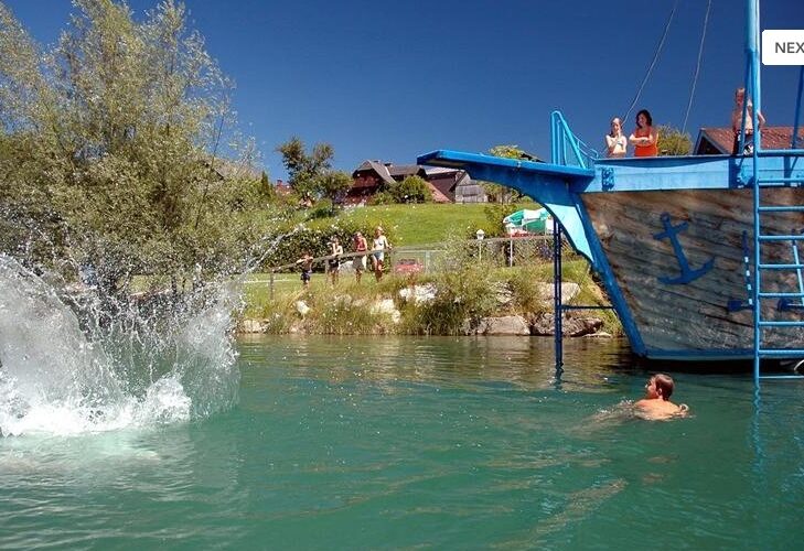 Die Leute schwimmen und sehen zu, wie jemand von einer blauen Holzkonstruktion in Form eines Schiffes in einen See springt und dabei einen großen Sprung macht.