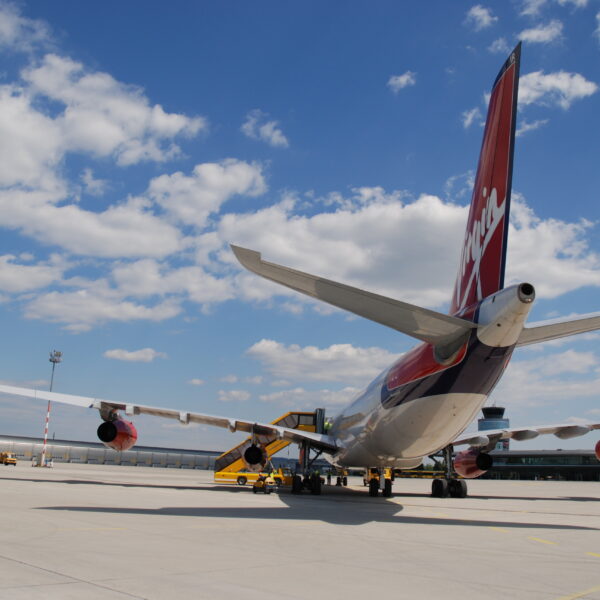 Ein großes Verkehrsflugzeug mit rotem Leitwerk steht bei teilweise bewölktem Himmel auf der Rollbahn, in der Nähe befinden sich Fahrzeuge des Bodenservice.