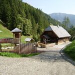 Ein Holzhaus und ein eingezäunter Spielplatz liegen an einer Schotterstraße, umgeben von bewaldeten Hügeln und fernen Bergen unter einem klaren Himmel.