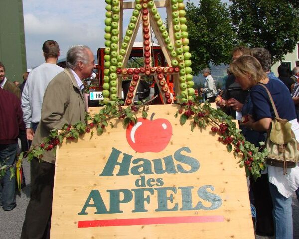 Menschen stehen bei einer Veranstaltung im Freien neben einem Holzschild mit der Aufschrift "Haus des Apfels", im Hintergrund eine Apfelauslage aus gestapelten Äpfeln.