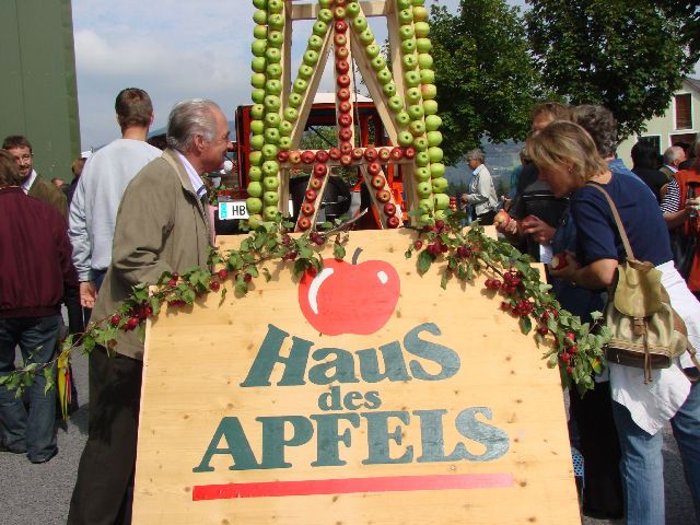 Menschen stehen bei einer Veranstaltung im Freien neben einem Holzschild mit der Aufschrift "Haus des Apfels", im Hintergrund eine Apfelauslage aus gestapelten Äpfeln.