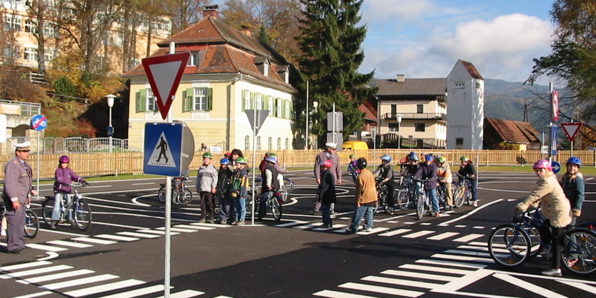 Eine Gruppe von Kindern mit Fahrrädern und Erwachsenen steht an einem Zebrastreifen in einer simulierten Straßenumgebung mit Verkehrsschildern und Straßenmarkierungen.