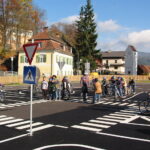 Eine Gruppe von Kindern mit Fahrrädern und Erwachsenen steht an einem Zebrastreifen in einer simulierten Straßenumgebung mit Verkehrsschildern und Straßenmarkierungen.