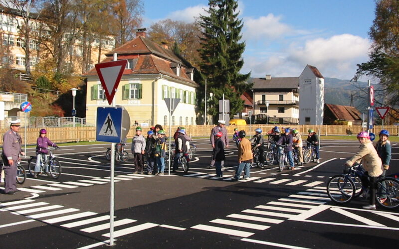 Eine Gruppe von Kindern mit Fahrrädern und Erwachsenen steht an einem Zebrastreifen in einer simulierten Straßenumgebung mit Verkehrsschildern und Straßenmarkierungen.