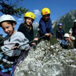 Eine Gruppe von Kindern und ein Erwachsener, die alle Helme und Klettergurte tragen, klettern auf einen Felsen in einer bergigen Umgebung.