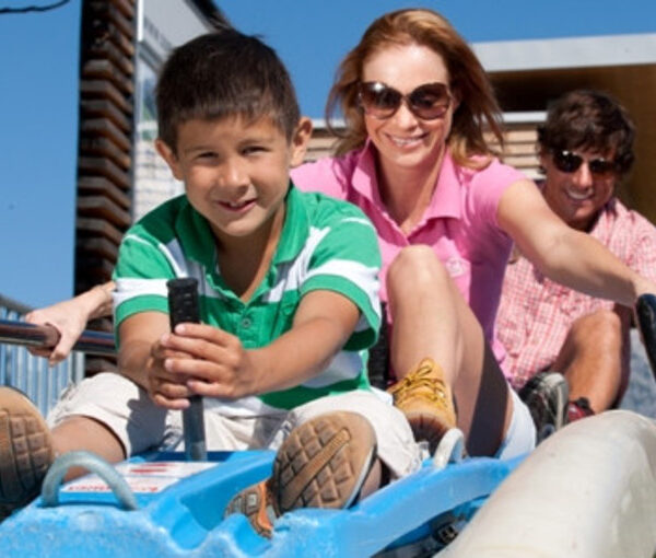 Ein kleiner Junge, eine Frau und ein Mann fahren gemeinsam auf einer blauen Alpenrutsche unter einem sonnigen Himmel, alle lächeln und genießen die Aktivität.
