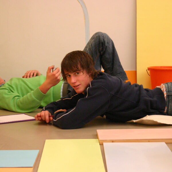 Zwei junge Menschen liegen in einem Raum mit bunten Oberflächen auf dem Boden, einer lächelt in die Kamera, der andere schreibt auf einem Klemmbrett. In der Nähe sind ein Spielzeuglastwagen und ein orangefarbener Eimer zu sehen.