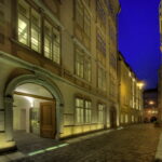 Eine schmale Kopfsteinpflasterstraße mit historischen Gebäuden unter einem tiefblauen Abendhimmel; ein Gebäude hat ein offenes Bogentor, dessen Eingang von Lichtern beleuchtet wird.