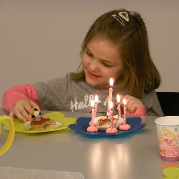 Ein junges Mädchen sitzt an einem Tisch mit einem Geburtstagsdessert mit Kerzen und einem Teller voller Essen, lächelt und greift nach einer Leckerei.