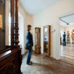 Eine Person geht durch einen Raum, in dem verschiedene antike Uhren ausgestellt sind, darunter große Standuhren und verzierte Holzgehäuse, in einer Museumsumgebung.