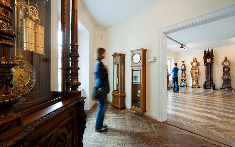 Eine Person geht durch einen Raum, in dem verschiedene antike Uhren ausgestellt sind, darunter große Standuhren und verzierte Holzgehäuse, in einer Museumsumgebung.