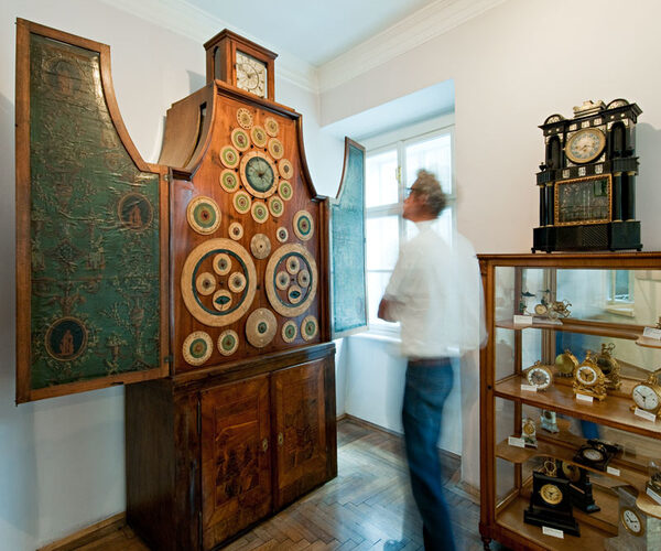 Eine Person steht neben einem großen, antiken Uhrenschrank mit mehreren Zifferblättern und offenen Türen, neben einer Vitrine mit kleineren Uhren in einem Museumsraum.