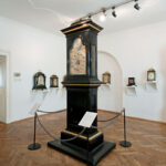 Ein Museumsraum mit verschiedenen antiken Uhren, darunter eine große schwarze Standuhr in der Mitte und mehrere andere Uhren, die in Regalen und an den Wänden ausgestellt sind.