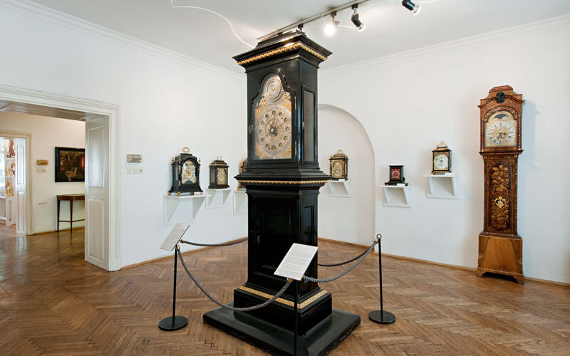 Ein Museumsraum mit verschiedenen antiken Uhren, darunter eine große schwarze Standuhr in der Mitte und mehrere andere Uhren, die in Regalen und an den Wänden ausgestellt sind.