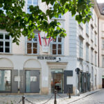 Straßenansicht des Wien Museum Uhrenmuseums, ein weißes Gebäude mit großen Fenstern, Beschilderung und mehreren Fahnen über dem Eingang in einer Kopfsteinpflasterstraße.