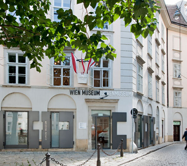 Außenansicht des Wien Museum Uhrenmuseums, mit großen Fenstern, Beschilderung und ein paar Menschen, die in der Nähe auf einer Kopfsteinpflasterstraße spazieren gehen.