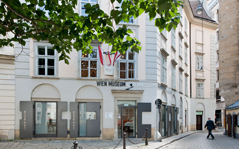 Außenansicht des Wien Museum Uhrenmuseums, mit großen Fenstern, Beschilderung und ein paar Menschen, die in der Nähe auf einer Kopfsteinpflasterstraße spazieren gehen.