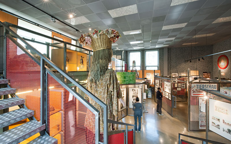 Innenansicht einer Museumsausstellung mit Schautafeln, gerahmten Gegenständen und einer großen Figur mit einer Blumenkrone und einem Korb auf dem Kopf neben einer Treppe.