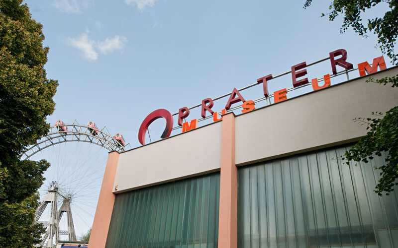 Gebäude des Pratermuseums mit Teilansicht des Wiener Riesenrads im Hintergrund, bei klarem Himmel und umgeben von Bäumen.