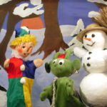 Drei Puppen - ein Clown, ein grünes Wesen und ein Schneemann mit einem Besen - stehen vor einer verschneiten Waldkulisse mit Bäumen und einer untergehenden Sonne.