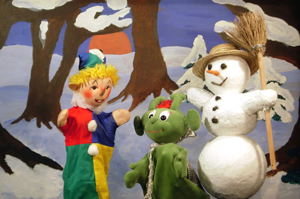 Drei Puppen - ein Clown, ein grünes Wesen und ein Schneemann mit einem Besen - stehen vor einer verschneiten Waldkulisse mit Bäumen und einer untergehenden Sonne.