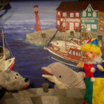 Drei Puppen - ein grauer Wolf, ein Delfin und ein Clown - treten vor einer gemalten Kulisse auf, die einen Hafen mit Booten und bunten Gebäuden zeigt.