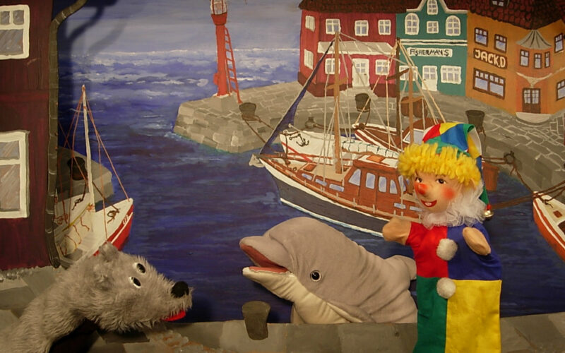 Drei Puppen - ein grauer Hund, ein Delfin und ein Clown - stehen vor einem gemalten Hintergrund, der einen Hafen mit Booten und bunten Gebäuden zeigt.
