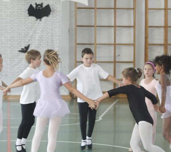 Eine Gruppe kleiner Kinder in Ballettkleidung hält sich in einer mit schwarzen Papierfledermäusen an den Wänden dekorierten Turnhalle im Kreis.
