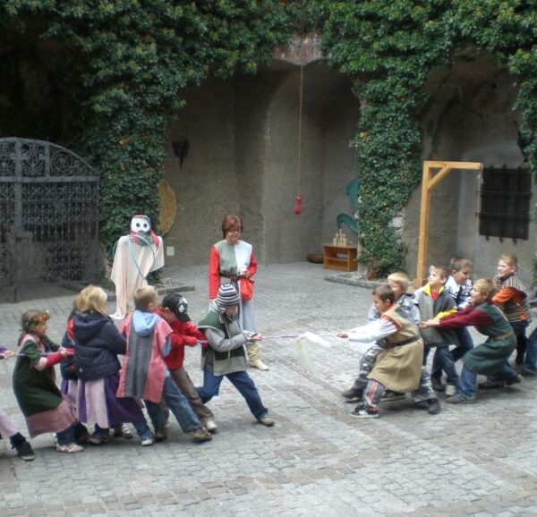 Eine Gruppe kostümierter Kinder spielt Tauziehen in einem gepflasterten Innenhof. Ein Erwachsener beaufsichtigt die Kinder und in der Nähe steht eine Person mit einer weißen Maske und einem Umhang.