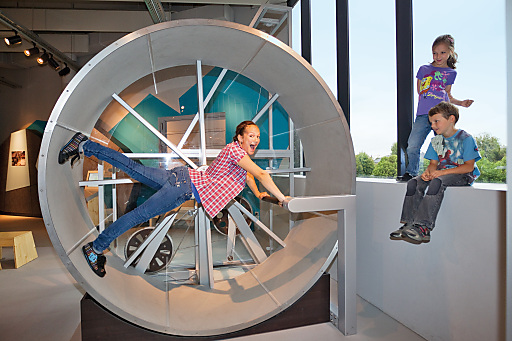 Ein Kind dreht sich in einem großen menschlichen Hamsterrad, während zwei andere Kinder in einem hell erleuchteten Innenraum von einem nahen Sims aus zusehen.