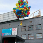 Eine Person in einem gelben Outfit schwebt über einer Menschenmenge, getragen von einer großen Gruppe bunter Luftballons vor einem modernen Gebäude.