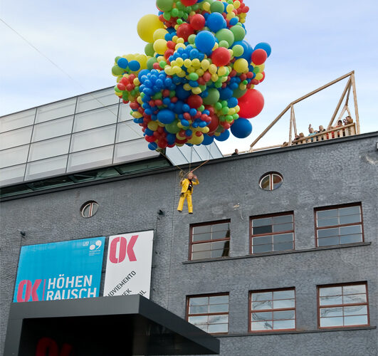 Eine Person in einem gelben Outfit schwebt über einer Menschenmenge, getragen von einer großen Gruppe bunter Luftballons vor einem modernen Gebäude.
