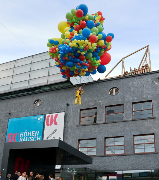Eine Person in einem gelben Outfit schwebt über einer Menschenmenge, getragen von einer großen Gruppe bunter Luftballons vor einem modernen Gebäude.