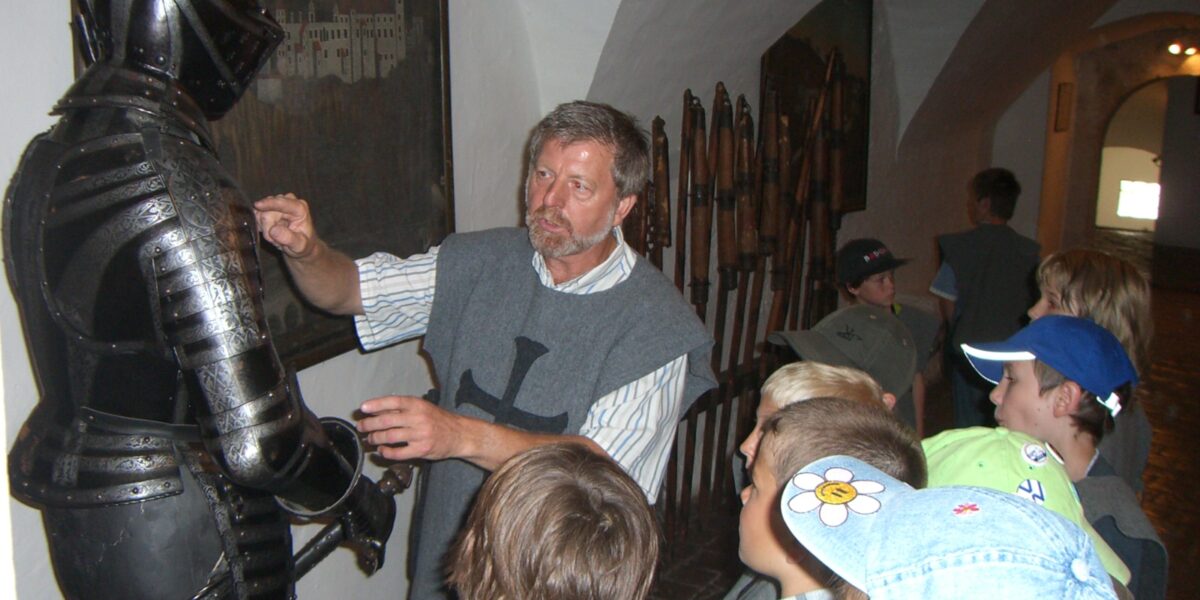 In einem Museumskorridor mit historischen Ausstellungsstücken an den Wänden erklärt ein Mann einer Gruppe von Kindern eine mittelalterliche Rüstung.