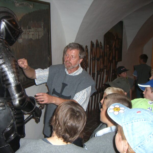 In einem Museumskorridor mit historischen Ausstellungsstücken an den Wänden erklärt ein Mann einer Gruppe von Kindern eine mittelalterliche Rüstung.
