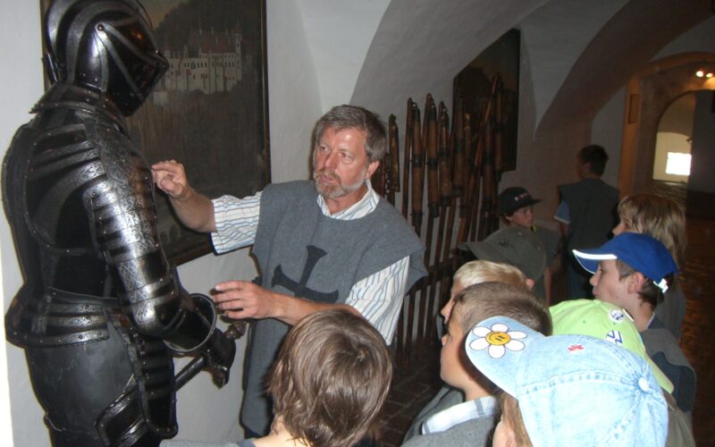 In einem Museumskorridor mit historischen Ausstellungsstücken an den Wänden erklärt ein Mann einer Gruppe von Kindern eine mittelalterliche Rüstung.