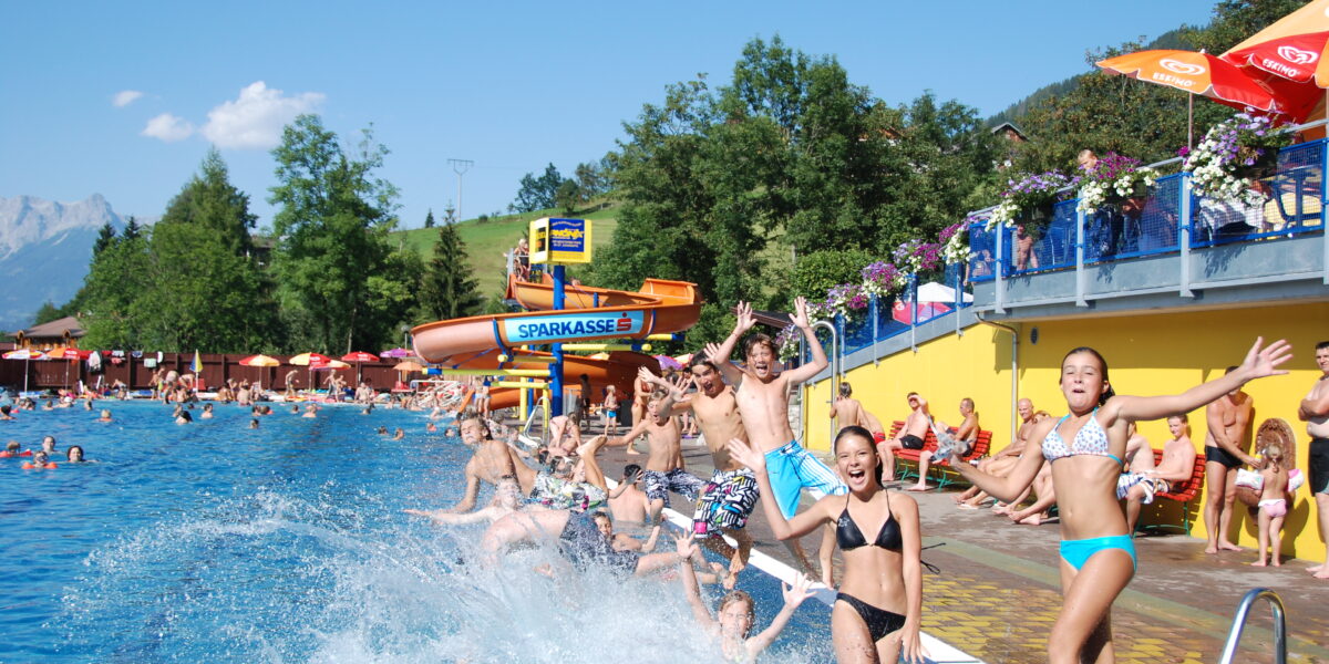 Eine Gruppe von Kindern und Jugendlichen springt an einem sonnigen Tag in ein Freibad, während sich die Leute am Beckenrand entspannen und eine Wasserrutsche im Hintergrund zu sehen ist.