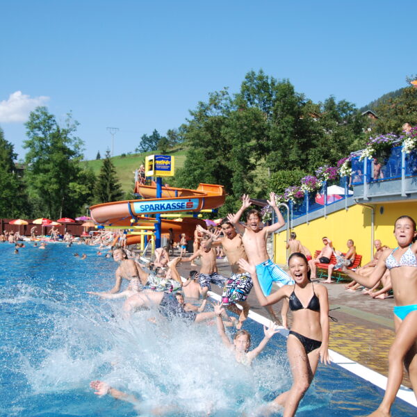 Eine Gruppe von Kindern und Jugendlichen springt an einem sonnigen Tag in ein Freibad, während sich die Leute am Beckenrand entspannen und eine Wasserrutsche im Hintergrund zu sehen ist.